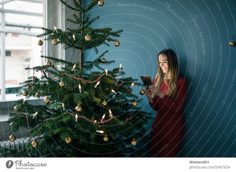 Smiling woman standing besides decorated Christmas tree using tablet females women digitizer Tablet Computer Tablet PC Tablet Computers iPad Digital Tablet