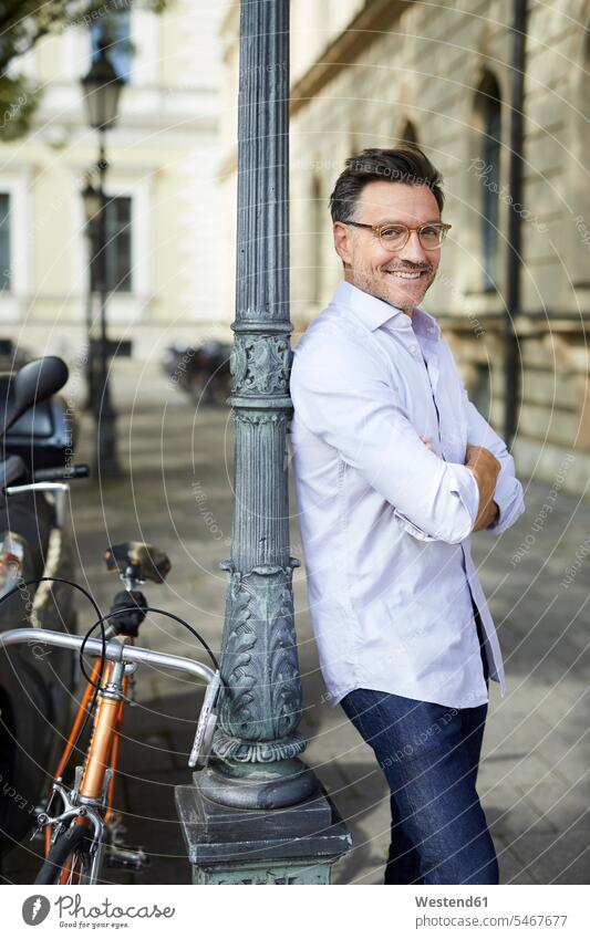 Portrait of smiling businessman with bicycle leaning against lamp post in the city portrait portraits smile town cities towns Businessman Business man