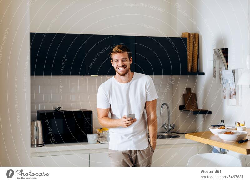 Portrait of happy young man holding cell phone in kitchen human human being human beings humans person persons celibate celibates singles solitary people