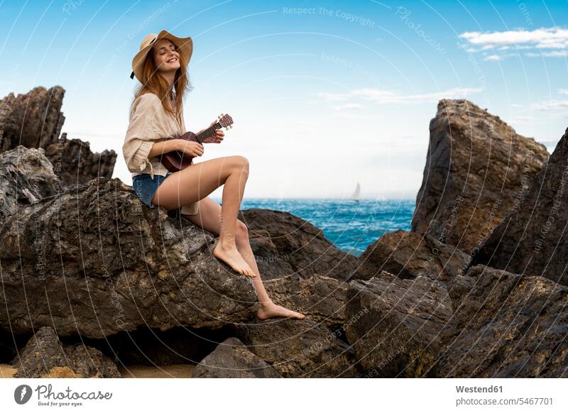 Happy young woman sitting on rock playing ukulele Spain water's edge waterside shore making music playing music make music play music carefree beauty beautiful