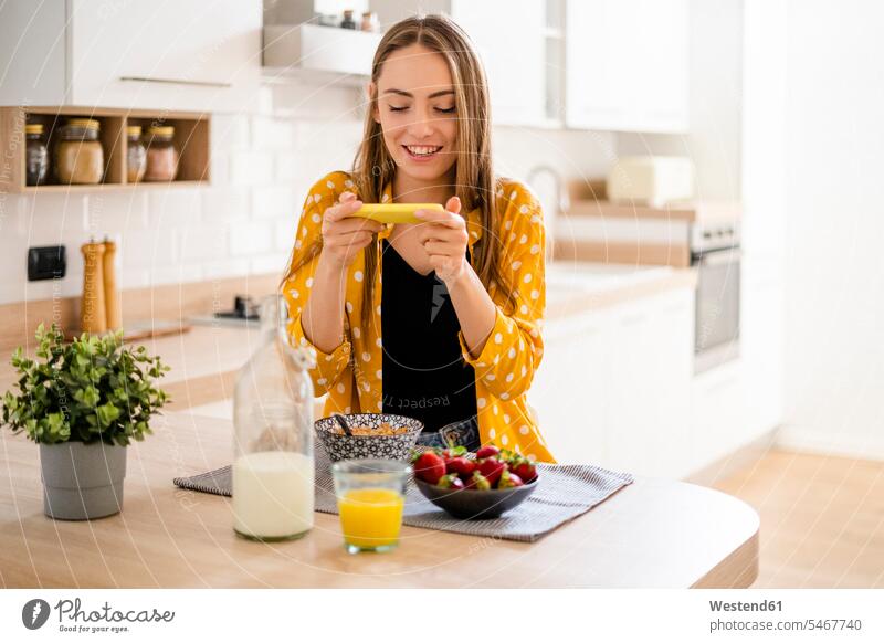 Happy young woman using cell phone and having breakfast in kitchen at home human human being human beings humans person persons celibate celibates singles