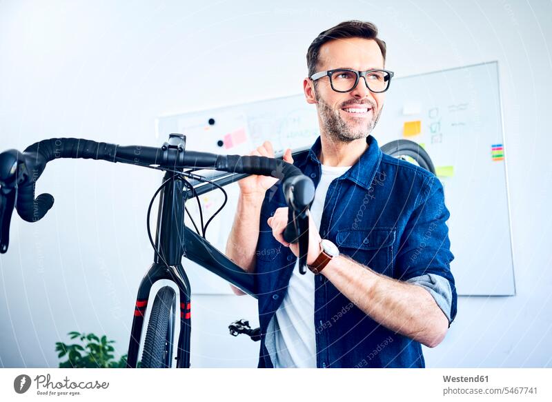 Smiling businessman carrying bicycle in office smiling smile bikes bicycles offices office room office rooms workplace work place place of work glasses specs