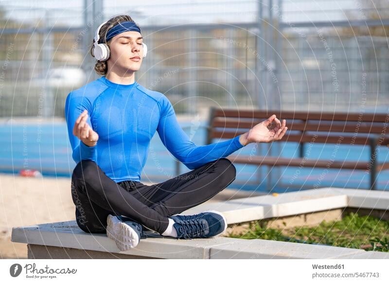 Young sportsman listening music while sitting in lotus position on retaining wall during sunny day color image colour image outdoors location shots outdoor shot