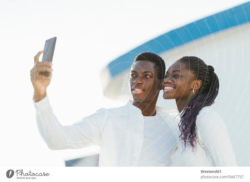 Smiling couple taking selfie through mobile phone against white wall color image colour image outdoors location shots outdoor shot outdoor shots day
