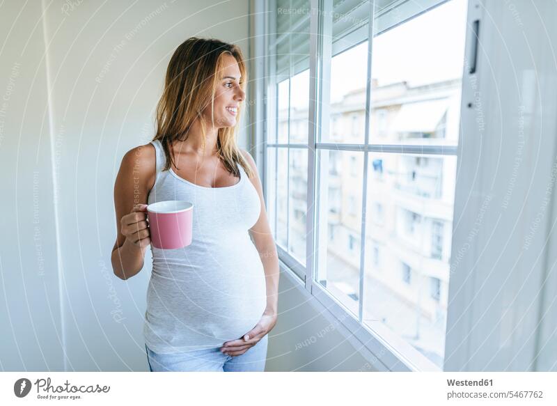 Pregnant woman looking out of window holding mug mugs cup pregnant Pregnant Woman windows females women Adults grown-ups grownups adult people persons