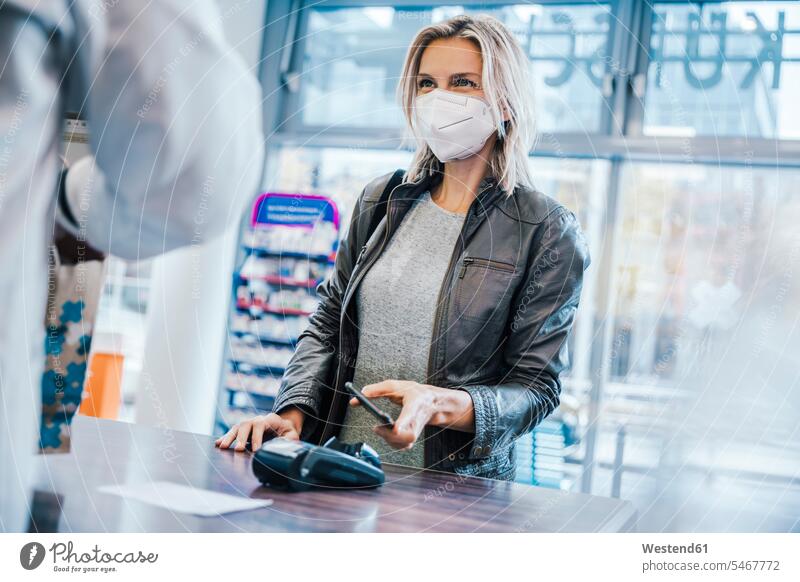 Female customer wearing face mask while making payment through smart phone at checkout counter in chemist shop color image colour image indoors indoor shot