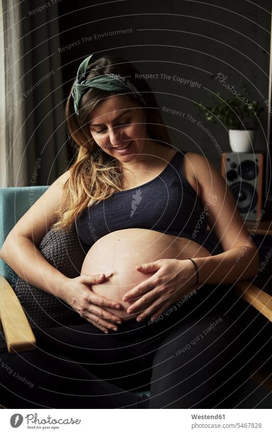 Pregnant woman looking on her baby belly at home chairs touch smile Seated sit delight enjoyment Pleasant pleasure happy caressing loving tender pleased