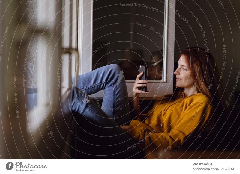 Smiling redheaded woman sitting at open window with feet up looking at cell phone human human being human beings humans person persons celibate celibates