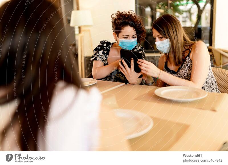 Women wearing masks while sharing digital tablet sitting at table in restaurant color image colour image Spain indoors indoor shot indoor shots interior