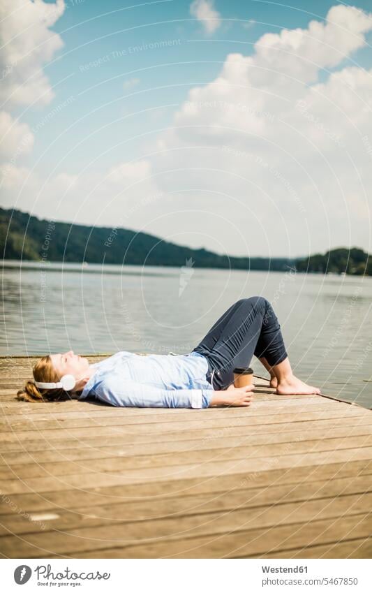 Woman lying on jetty at a lake with headphones and takeaway coffee headset jetties Coffee laying down lie lying down woman females women lakes Drink beverages