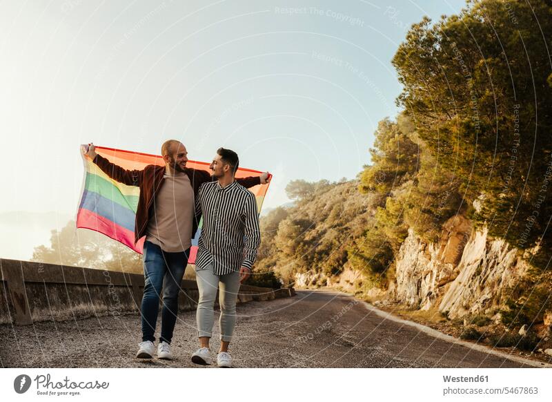 Gay couple with gay pride flag walking on a road in the mountains flags banner banners go going smile speak speaking talk delight enjoyment Pleasant pleasure