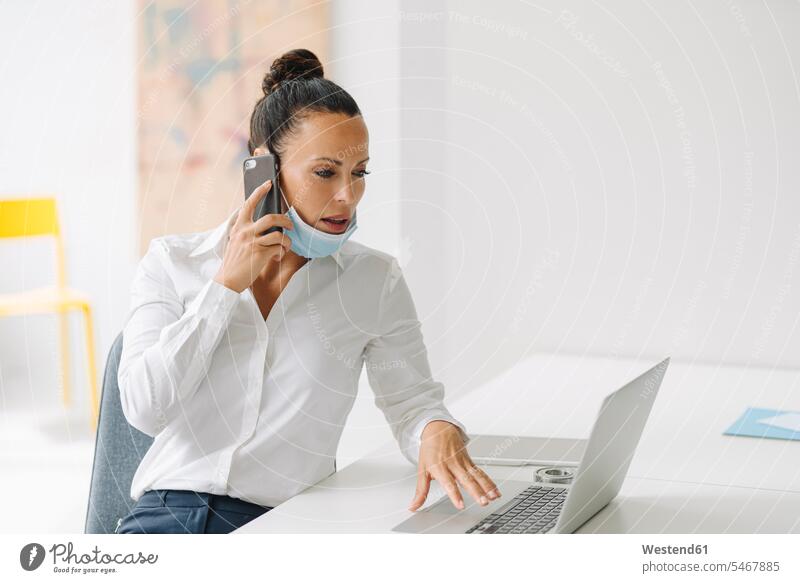 Businesswoman wearing mask talking over smart phone while using laptop on desk in home office color image colour image Germany indoors indoor shot indoor shots