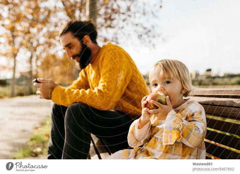 Father and daughter sitting on a bench in the park in autumn, father using smartphone, daughter eating an apple Apple Apples use Smartphone iPhone Smartphones