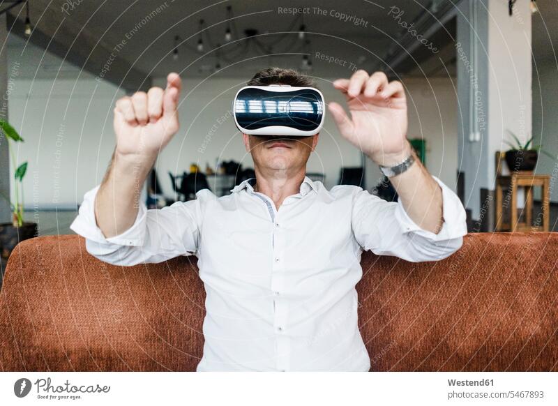 Mature man wearing VR glasses sitting on couch in a loft lofts Seated virtual reality specs Eye Glasses spectacles Eyeglasses men males settee sofa sofas