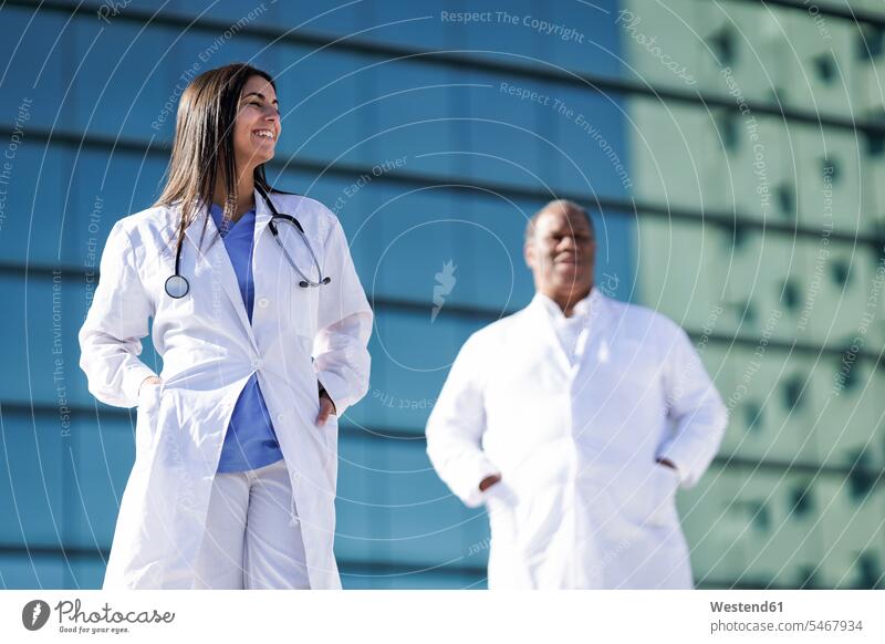 Confident doctors standing with hands in pockets against hospital color image colour image outdoors location shots outdoor shot outdoor shots day daylight shot