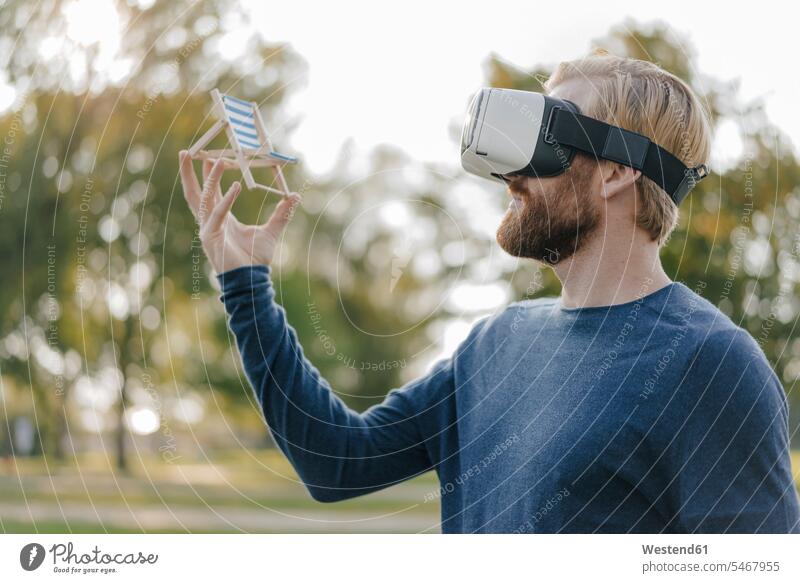 Man in autumnal park wearing Virtual Reality Glasses looking at miniature beach lounger beach loungers man men males VR glasses Virtual-Reality Glasses