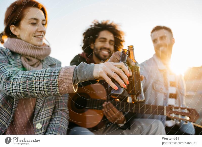 Three happy friends with guitar toasting beer bottles at sunset sitting Seated sunsets sundown clinking cheers happiness Beer Bottle Beer Bottles guitars
