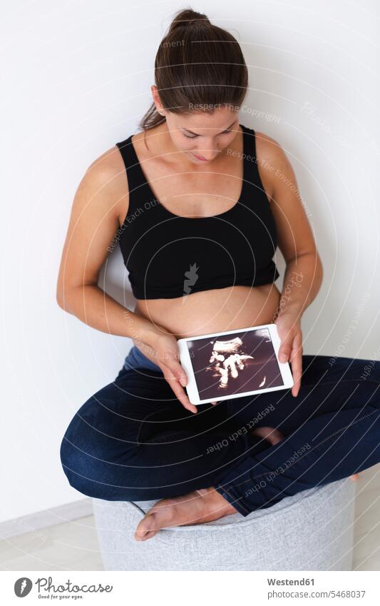 Young pregnant woman with ultrasound image on her tablet of her unborn baby upper part of the body bellies stomach stomachs hold Seated sit delight enjoyment