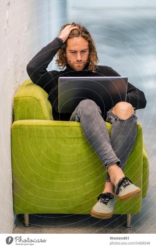 Young man with hand in hair using laptop while sitting on armchair at home color image colour image Spain leisure activity leisure activities free time