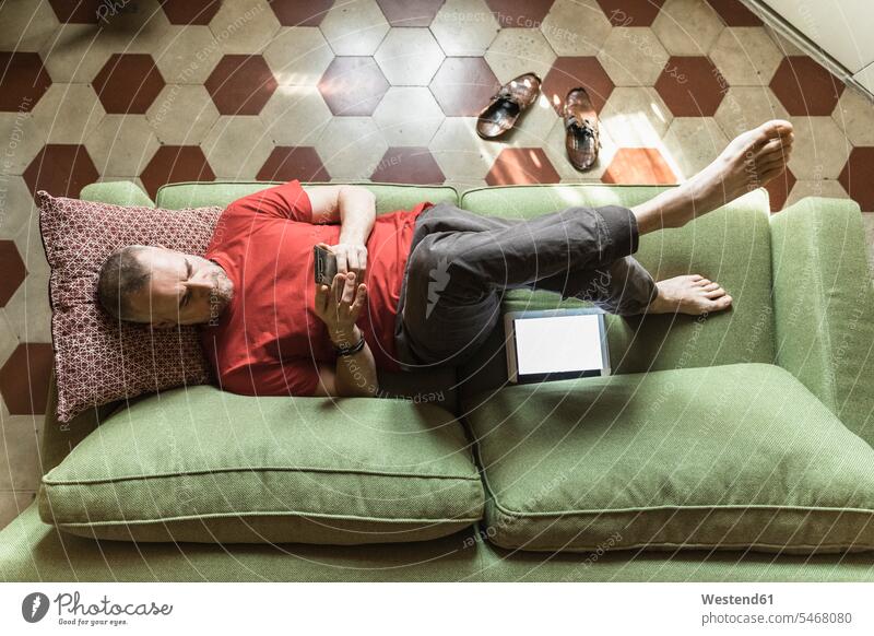 Mature man lying on sofa in living room and using tablet cushions couches settee settees sofas telecommunication phones telephone telephones cell phone
