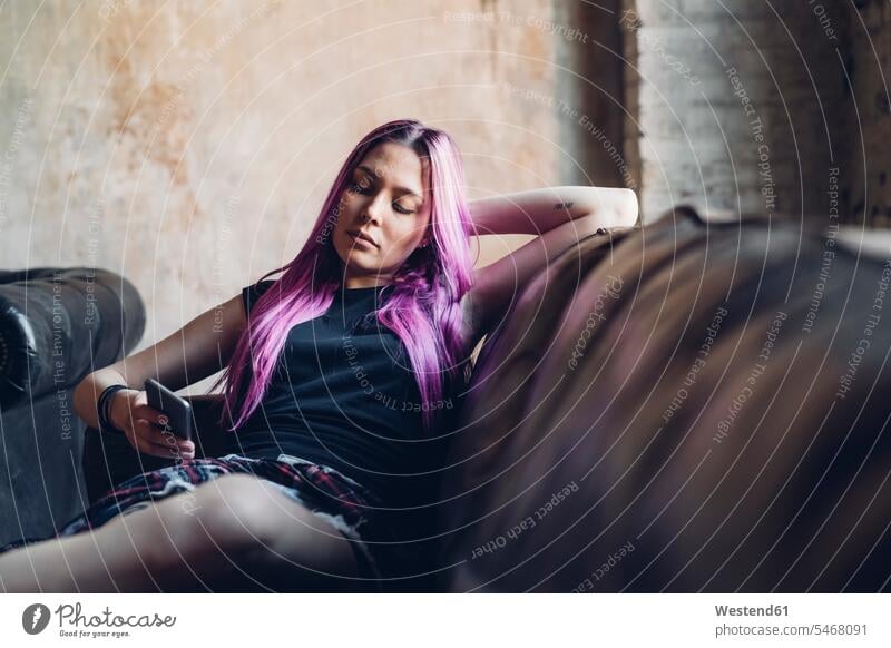 Young woman with pink hair sitting on sofa in a loft using smartphone couches settee settees sofas Seated relax relaxing relaxation colour colours magenta