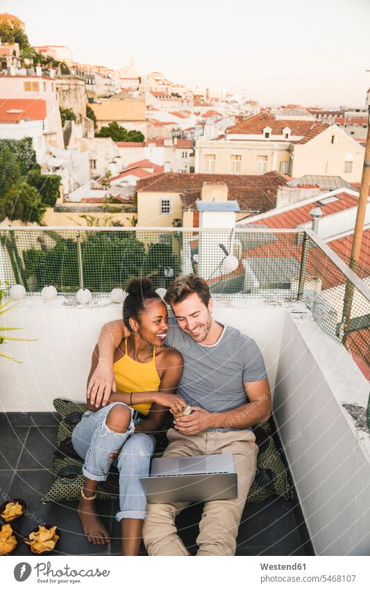 Happy young couple with laptop sitting on rooftop in the evening, Lisbon, Portugal cushions computers Laptop Computer Laptop Computers laptops notebook relax