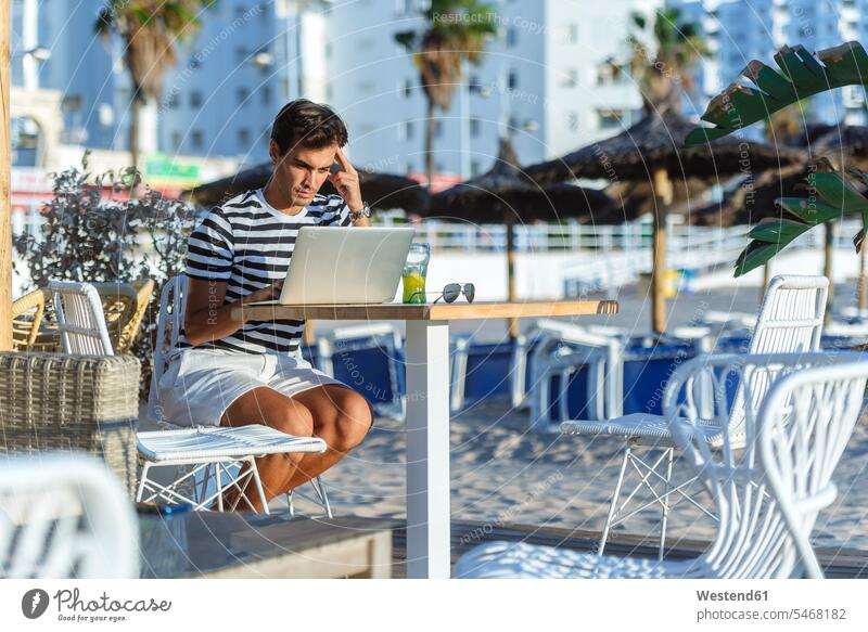 Man with laptop sitting on the terrace of a beach bar Seated Laptop Computers laptops notebook terraces man men males beaches bars computer computers Adults