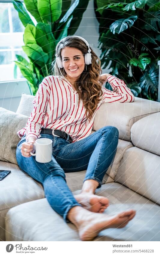 Portrait of smiling woman with cup of coffee and headphones sitting on couch Coffee portrait portraits females women Seated Coffee Cup Coffee Cups headset