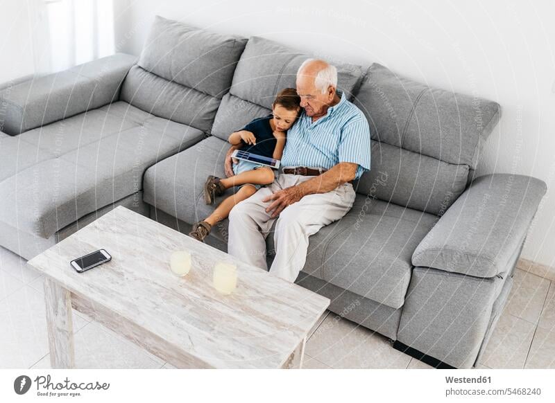Grandfather and grandson sitting together on the couch at home looking at digital tablet grandfather grandpas granddads grandfathers settee sofa sofas couches