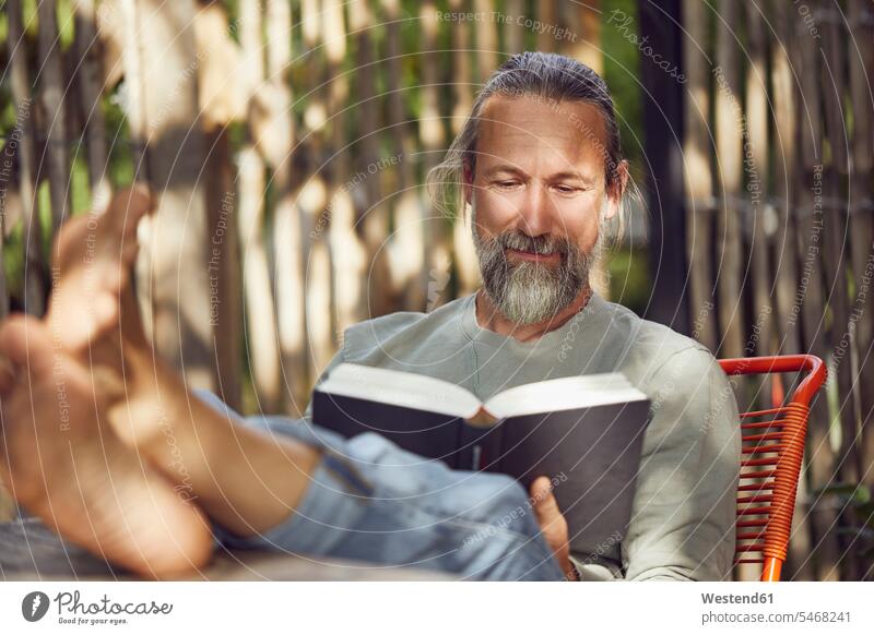 Bearded mature man reading book while relaxing on chair in yard color image colour image Germany leisure activity leisure activities free time leisure time