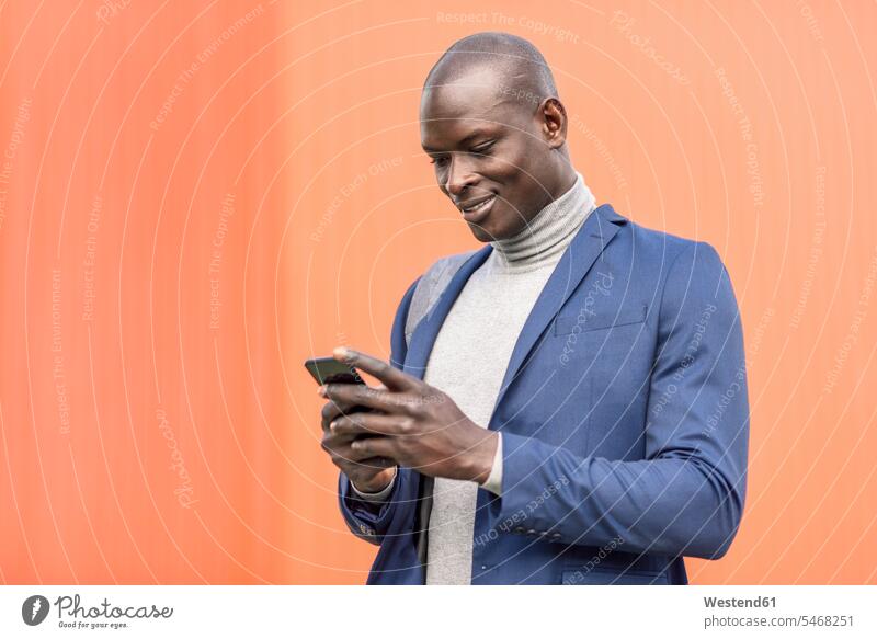 Smiling businessman standing in front of orange wall looking at smartphone Smartphone iPhone Smartphones walls eyeing portrait portraits Businessman