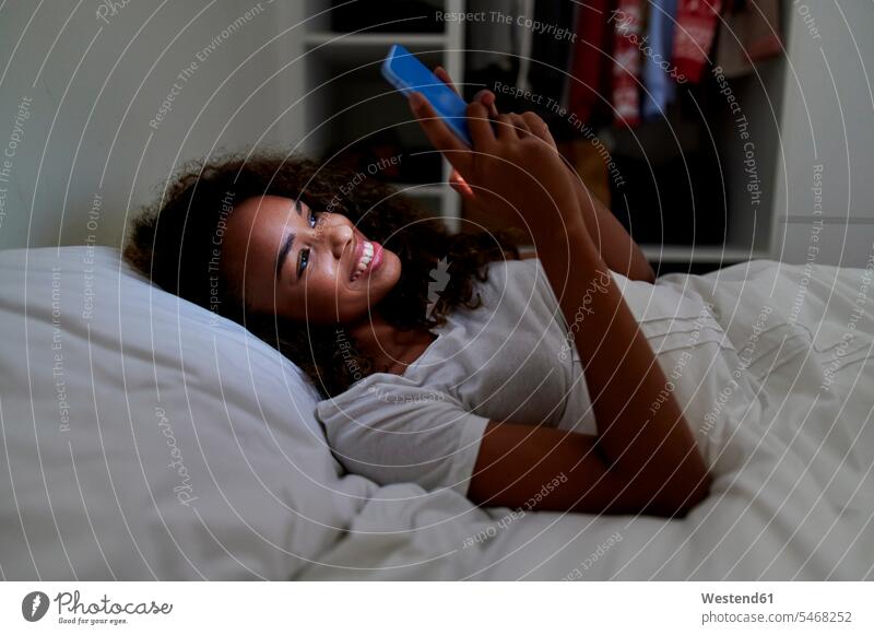 Happy young woman using smart phone while lying on bed in bedroom at home color image colour image indoors indoor shot indoor shots interior interior view