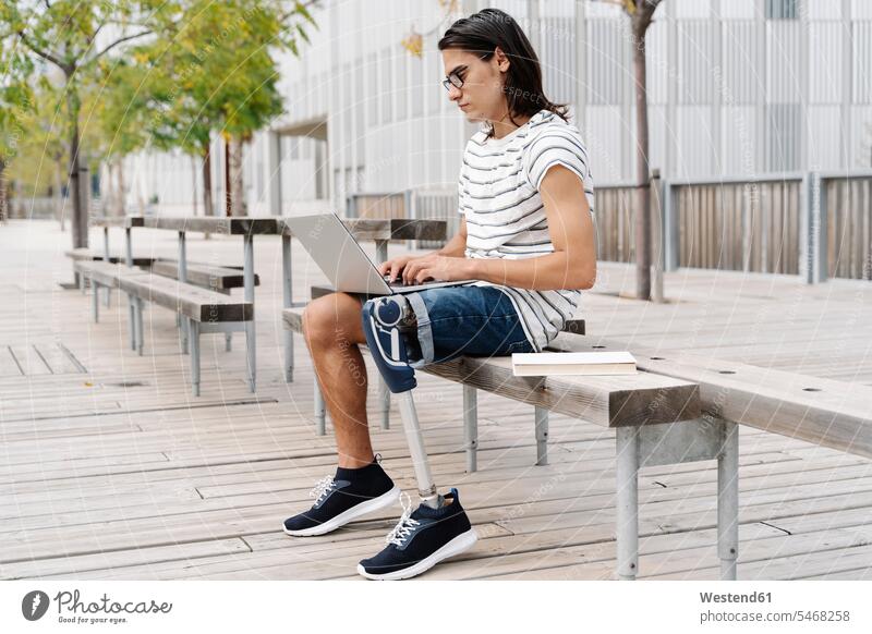 Contemplating man with artificial limb using laptop while sitting on bench in city color image colour image Spain outdoors location shots outdoor shot