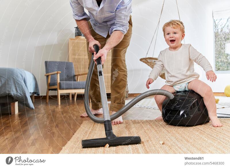Low section of man cleaning carpet while cute baby boy sitting on vacuum cleaner in living room at home color image colour image Germany indoors indoor shot