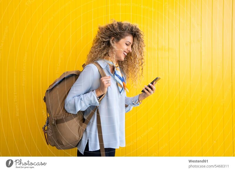 Smiling young woman with backpack in front of yellow background looking at cell phone human human being human beings humans person persons caucasian appearance