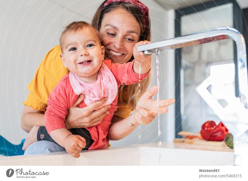Smiling mother and cute baby girl playing with water falling from faucet in kitchen sink color image colour image indoors indoor shot indoor shots interior
