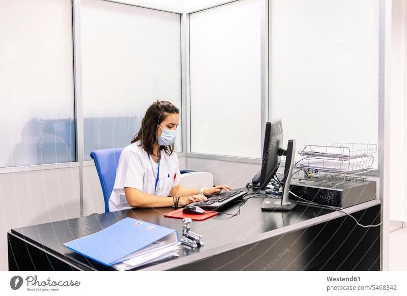 Female doctor wearing surgical mask using computer on desk in office at hospital color image colour image Spain indoors indoor shot indoor shots interior