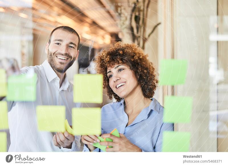 Two happy colleagues brainstorming with post-its on glass pane smiling smile happiness glass panes Brainstorming Adhesive Note post-it note sticky notes