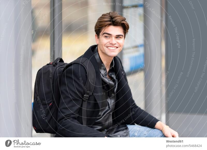 Smiling handsome young man looking away while sitting with backpack at bus stop color image colour image Spain outdoors location shots outdoor shot