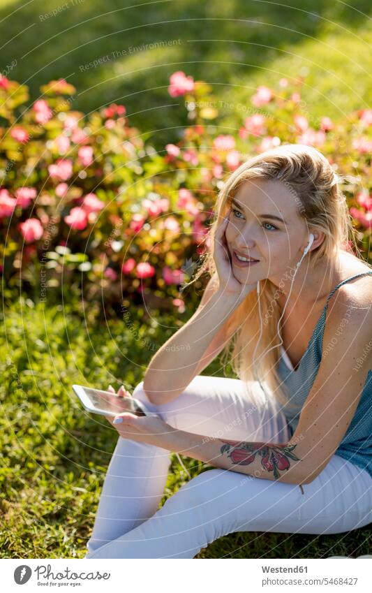 Smiling young woman sitting in park with cell phone and earbuds mobile phone mobiles mobile phones Cellphone cell phones earphones ear phone ear phones females