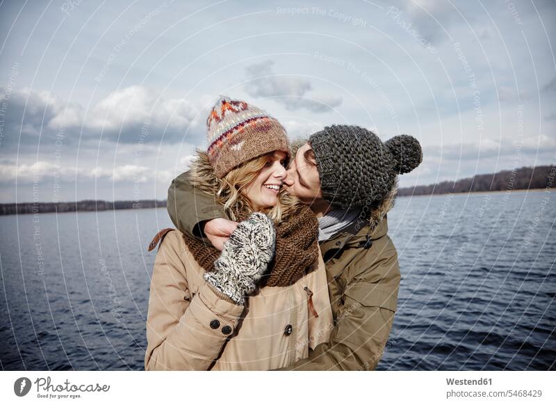 Young man kissing his girlfriend in front of a lake gloves kisses seasons hibernal happy Emotions Feeling Feelings Sentiment Sentiments loving closeness