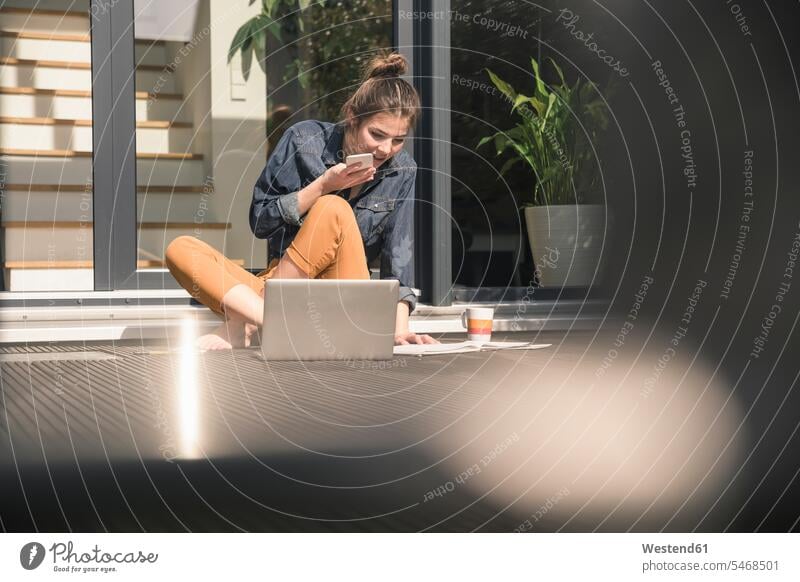 Young woman sitting on terrace at home using cell phone and laptop Germany freelancer freelancing working from home working at home Work From Home