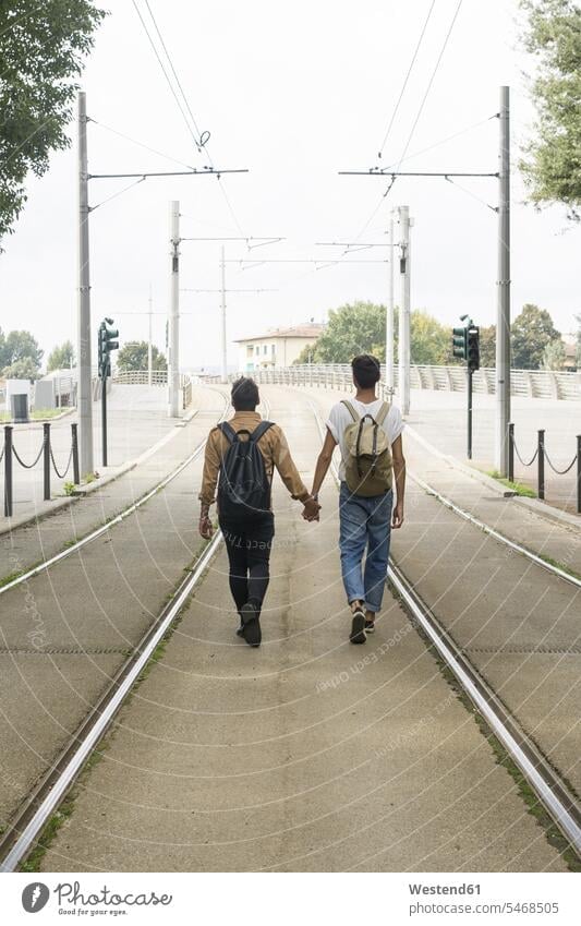 Back view of young gay couple with backpacks walking hand in hand between tracks on the road going twosomes partnership couples gay men gay man homosexual men