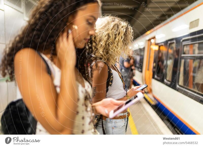 UK, London, two young women with cell phones waiting at underground station platform female friends woman females mobile phone mobiles mobile phones Cellphone