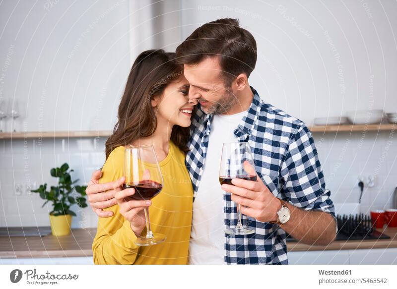 Happy couple standing in kitchen, with arms around, drinking red wine, kissing kisses domestic kitchen kitchens affectionate tender loving caressing happiness