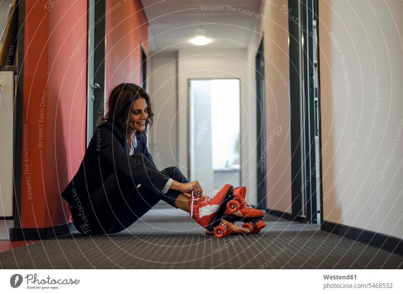 Mature businesswoman sitting in office corridor, putting on roller skates offices office room office rooms Seated businesswomen business woman business women