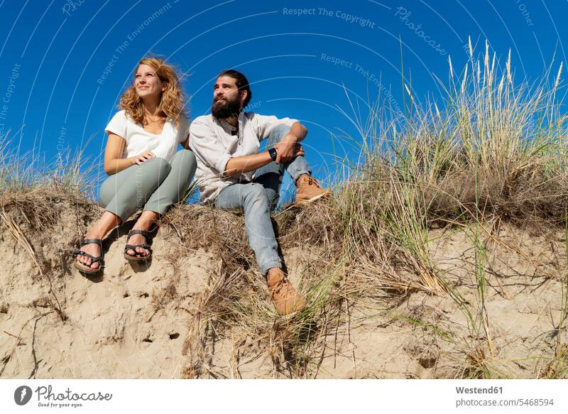 Young couple sitting on a dune in summer, relaxing Seated recovering young couple young couples young twosome young twosomes dunes summer time summery