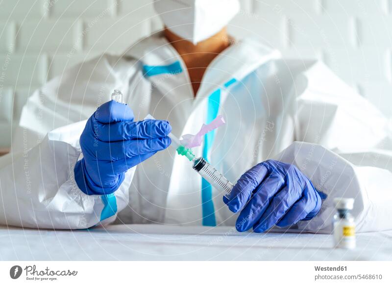 Nurse in protective workwear filling injection while sitting by table in hospital color image colour image indoors indoor shot indoor shots interior