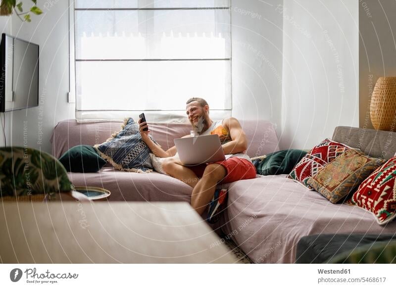 Smiling man using smart phone while sitting with laptop on sofa in living room color image colour image indoors indoor shot indoor shots interior interior view