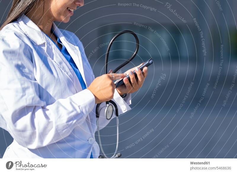 Doctor with stethoscope using mobile phone while standing against hospital color image colour image outdoors location shots outdoor shot outdoor shots day
